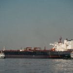 Navio cargueiro atracado em um porto, com rebocadores ao redor em um dia claro.