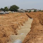 Canteiro de obras em área suburbana, com valas abertas e grandes tubulações instaladas sob céu claro.