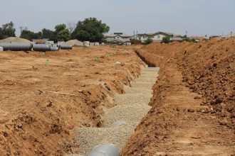 Canteiro de obras em área suburbana, com valas abertas e grandes tubulações instaladas sob céu claro.