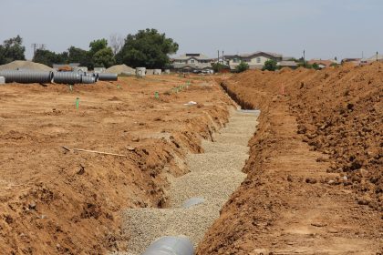 Canteiro de obras em área suburbana, com valas abertas e grandes tubulações instaladas sob céu claro.