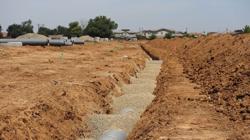 Canteiro de obras em área suburbana, com valas abertas e grandes tubulações instaladas sob céu claro.