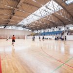 Atletas treinam em uma quadra de basquete coberta, em uma arena esportiva.