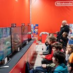 Pessoas jogam diante de várias telas em um evento de videogames, em ambiente iluminado e movimentado.