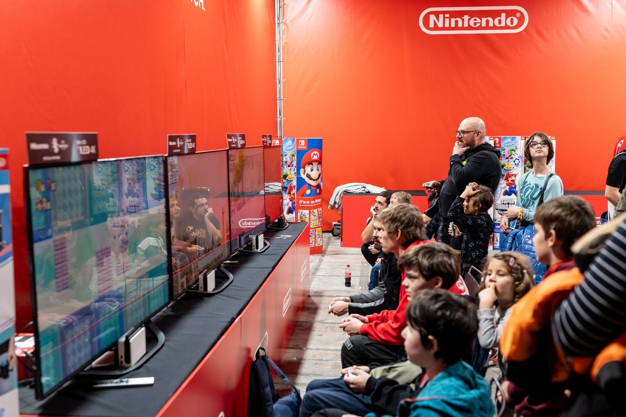 Pessoas jogam diante de várias telas em um evento de videogames, em ambiente iluminado e movimentado.