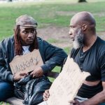 Dois homens sentados em um banco de parque seguram cartazes sobre fome e busca por trabalho.