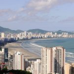 Vista aérea panorâmica de Santos, com edifícios altos junto à orla, o mar e morros ao fundo.