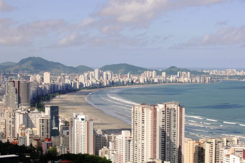 Vista aérea panorâmica de Santos, com edifícios altos junto à orla, o mar e morros ao fundo.